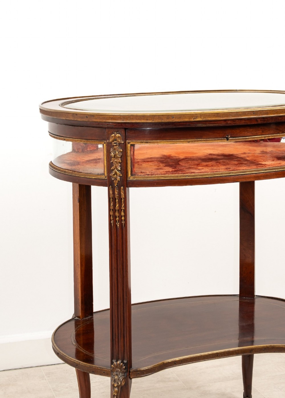 An antique oval mahogany bijouterie table featuring curved glass sides, an undertier shelf, and ornate gilt-bronze mounts. View VI