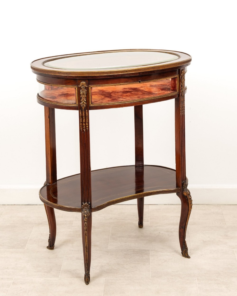 An antique oval mahogany bijouterie table featuring curved glass sides, an undertier shelf, and ornate gilt-bronze mounts. View VII