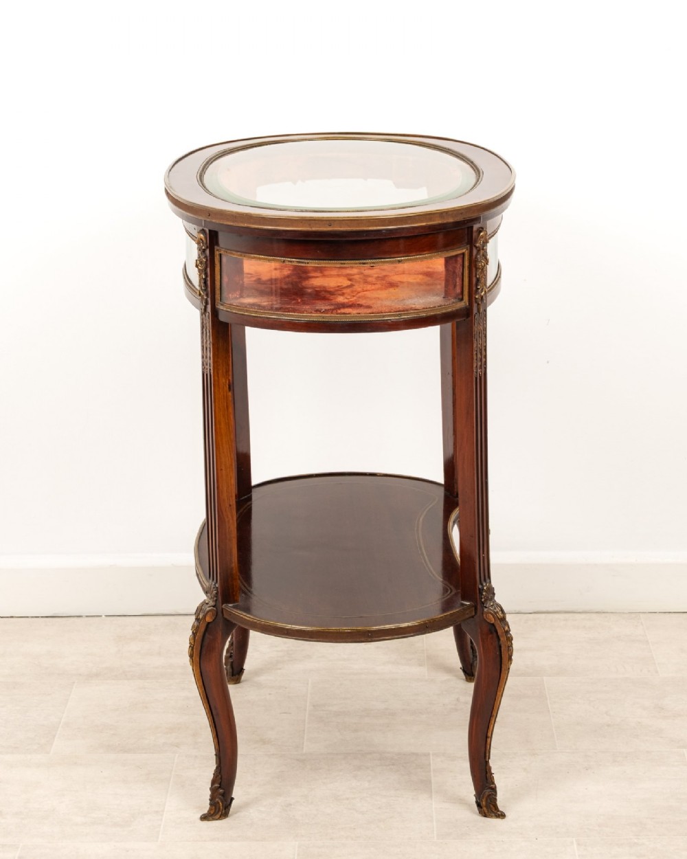 An antique oval mahogany bijouterie table featuring curved glass sides, an undertier shelf, and ornate gilt-bronze mounts. View VIII