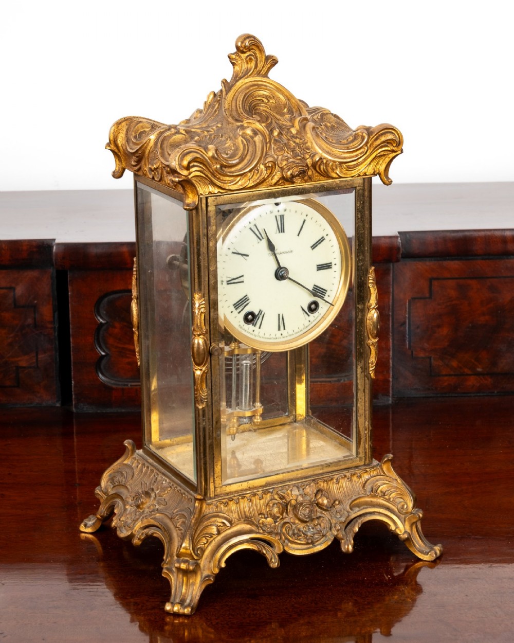 A late 19th-century Rococo Revival gilt bronze crystal regulator mantel clock featuring four bevelled glass panels, ornate scrollwork casting, an enamel dial with Roman numerals, and a visible mercury-style pendulum.