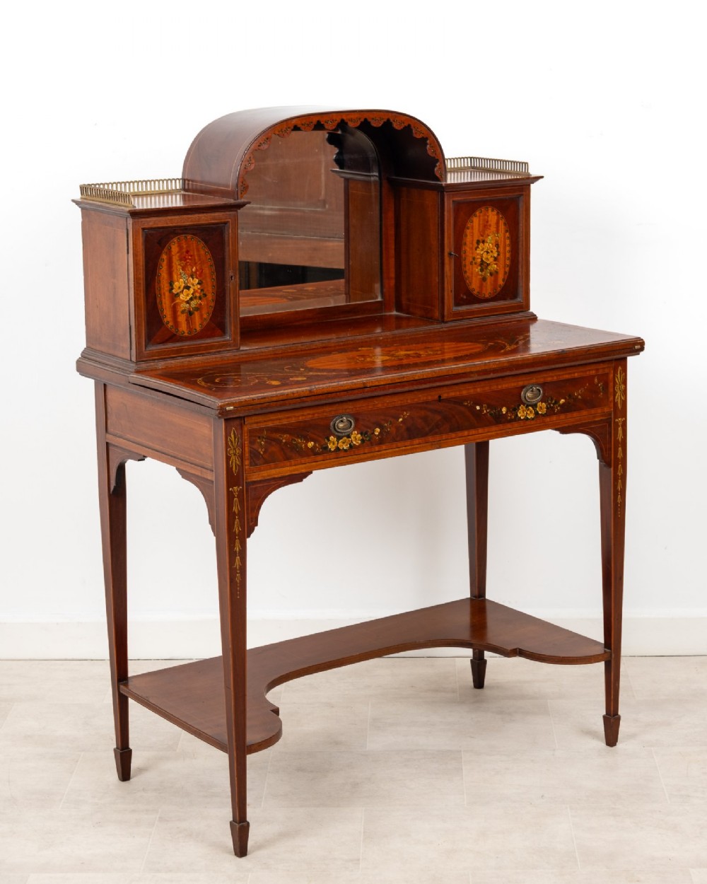 An Edwardian mahogany writing desk featuring floral hand-painted motifs, twin upper cabinets with brass galleries, an arched central mirror, and a single frieze drawer.