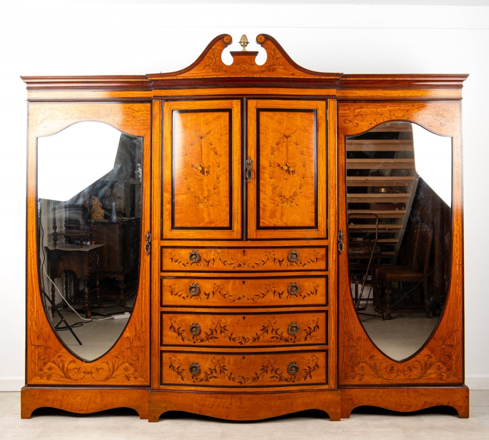 An Edwardian satinwood compactum wardrobe featuring twin arched mirror doors flanking a central cupboard and bank of four drawers, topped with a scroll pediment.