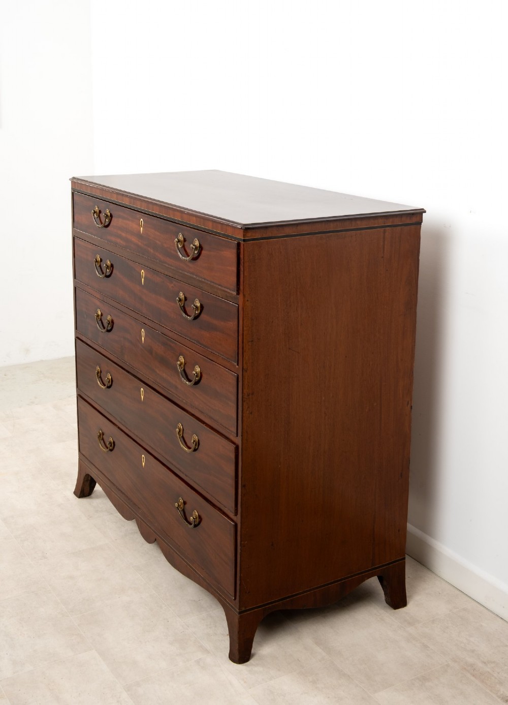 A nineteenth-century mahogany chest of five drawers featuring brass drop handles, keyhole escutcheons, and a shaped lower apron. View VI