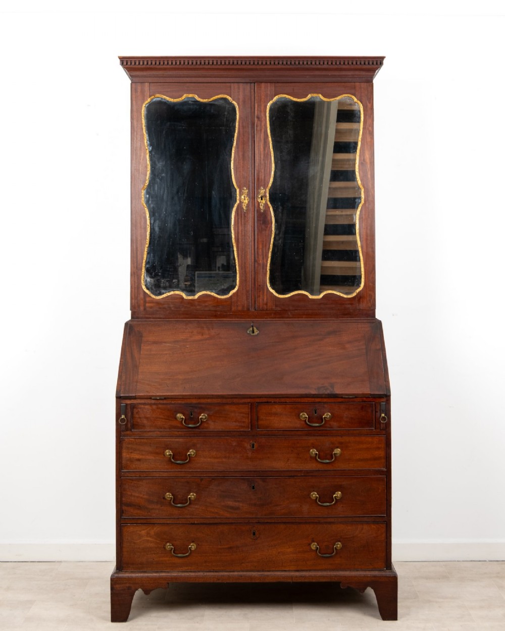 An eighteenth-century mahogany bureau bookcase featuring shaped mirrored double doors on the upper cabinet, a slant-front desk midsection, and four graduated drawers below with brass drop handles.