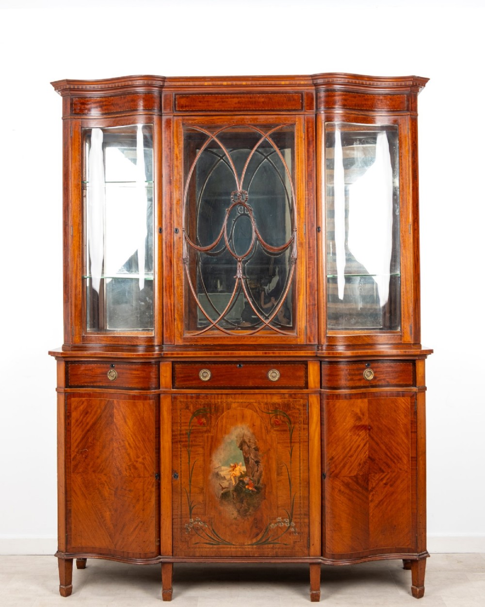 An Edwardian mahogany serpentine display cabinet featuring a central glazed door with geometric astragal glazing, glass side panels, and a hand-painted floral landscape panel on the lower central door.