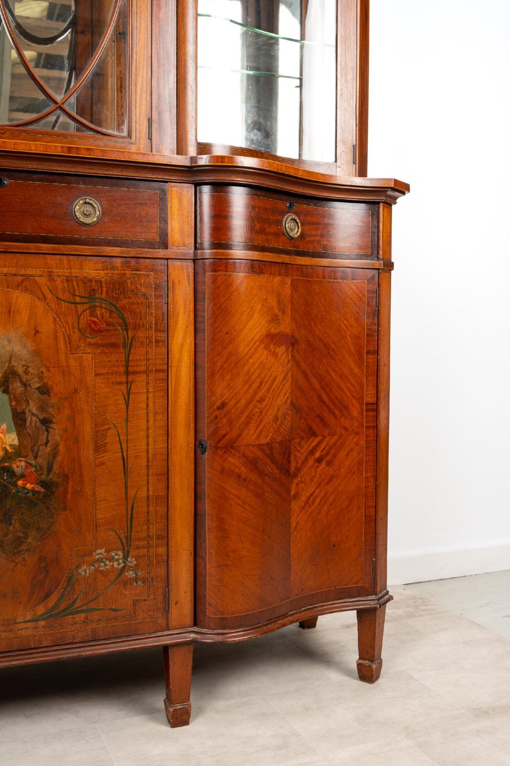 An Edwardian mahogany serpentine display cabinet featuring a central glazed door with geometric astragal glazing, glass side panels, and a hand-painted floral landscape panel on the lower central door. View III