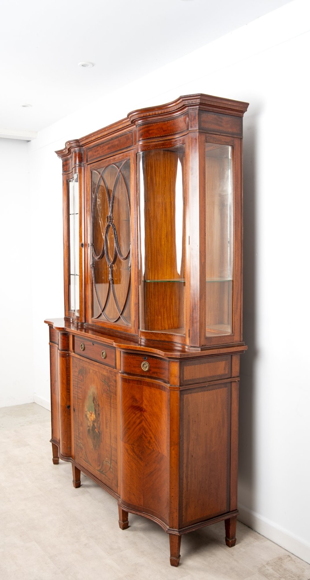 An Edwardian mahogany serpentine display cabinet featuring a central glazed door with geometric astragal glazing, glass side panels, and a hand-painted floral landscape panel on the lower central door. View VII