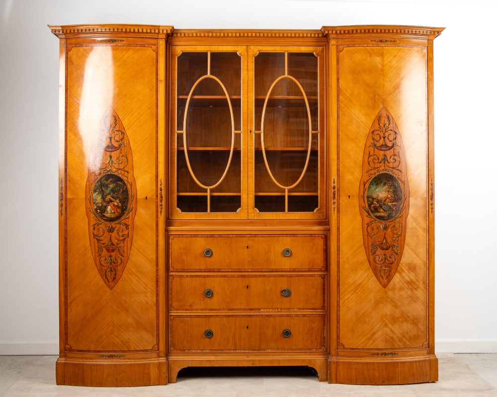 An Edwardian satinwood breakfront compactum wardrobe featuring central glazed doors, three drawers, and flanking bow-front cupboards decorated with hand-painted oval medallions.