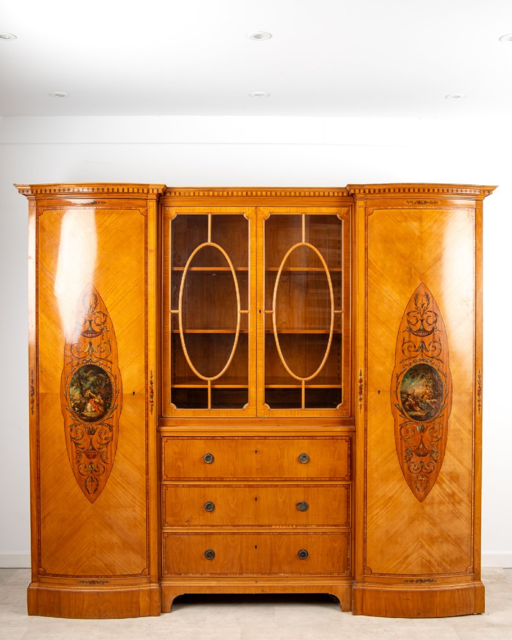 An Edwardian satinwood breakfront compactum wardrobe featuring central glazed doors, three drawers, and flanking bow-front cupboards decorated with hand-painted oval medallions. View III
