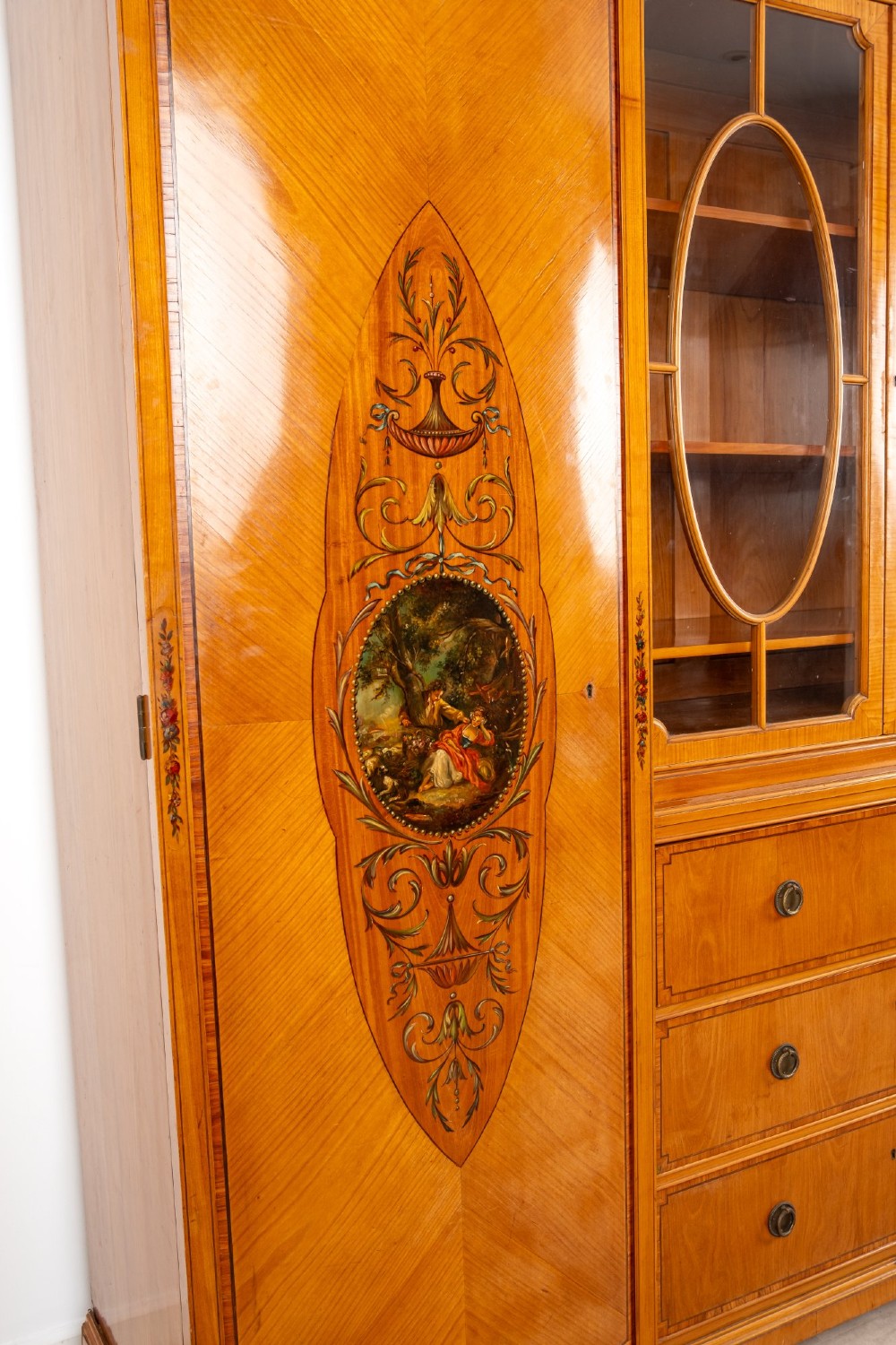 An Edwardian satinwood breakfront compactum wardrobe featuring central glazed doors, three drawers, and flanking bow-front cupboards decorated with hand-painted oval medallions. View IV