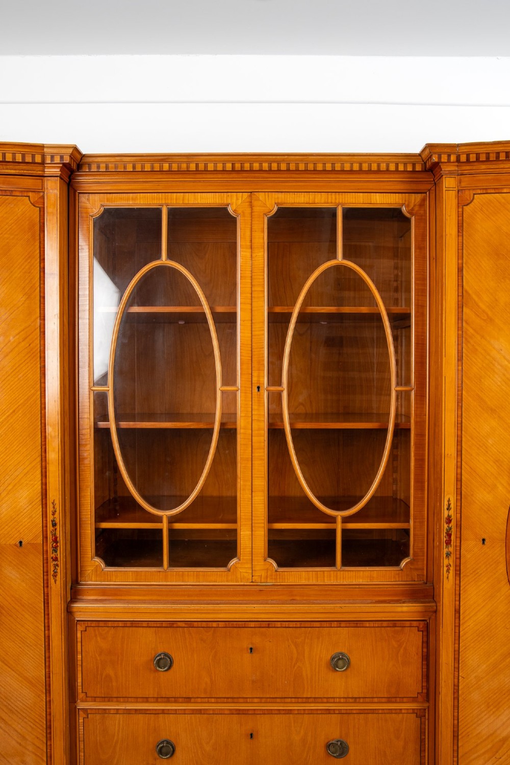 An Edwardian satinwood breakfront compactum wardrobe featuring central glazed doors, three drawers, and flanking bow-front cupboards decorated with hand-painted oval medallions. View V