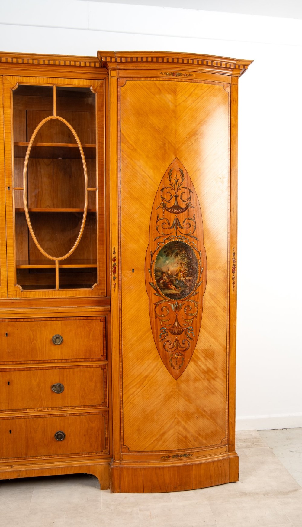 An Edwardian satinwood breakfront compactum wardrobe featuring central glazed doors, three drawers, and flanking bow-front cupboards decorated with hand-painted oval medallions. View VI