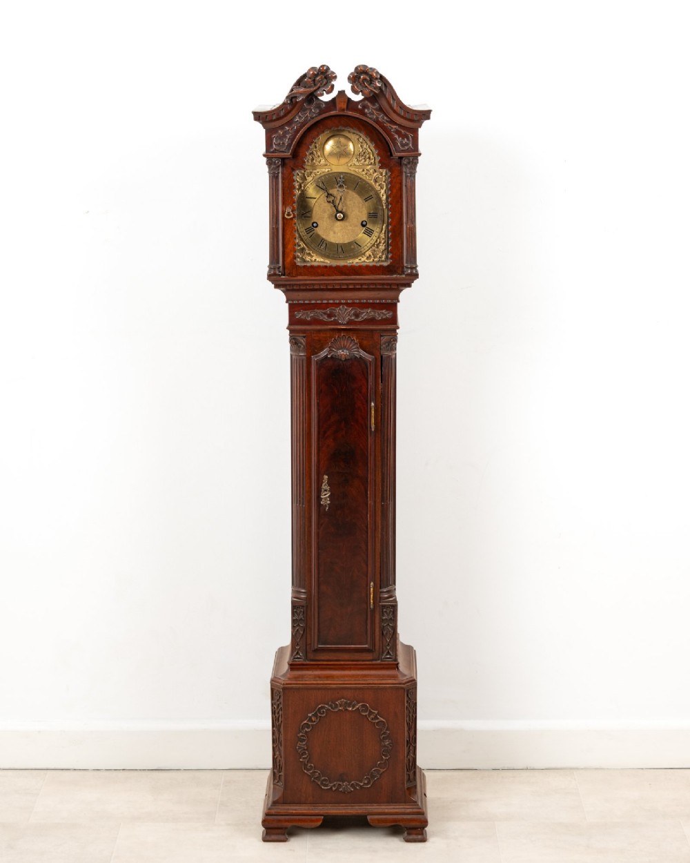An Edwardian mahogany grandmother clock with a carved swan neck pediment, a brass dial with Roman numerals, and a slim waist door. View II