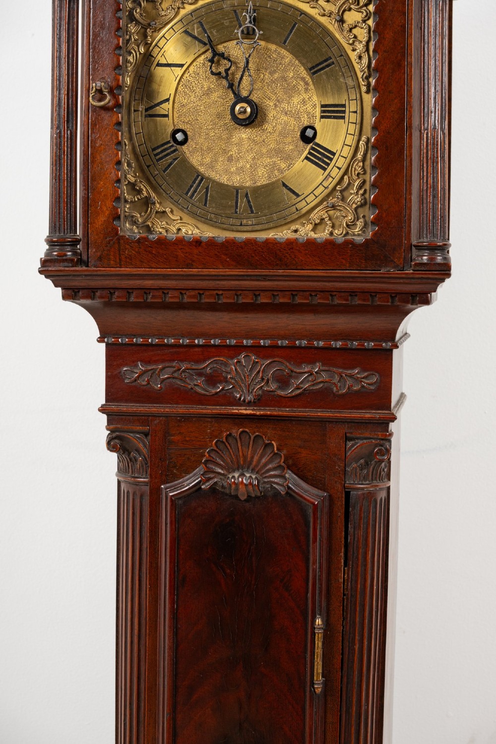 An Edwardian mahogany grandmother clock with a carved swan neck pediment, a brass dial with Roman numerals, and a slim waist door. View VI