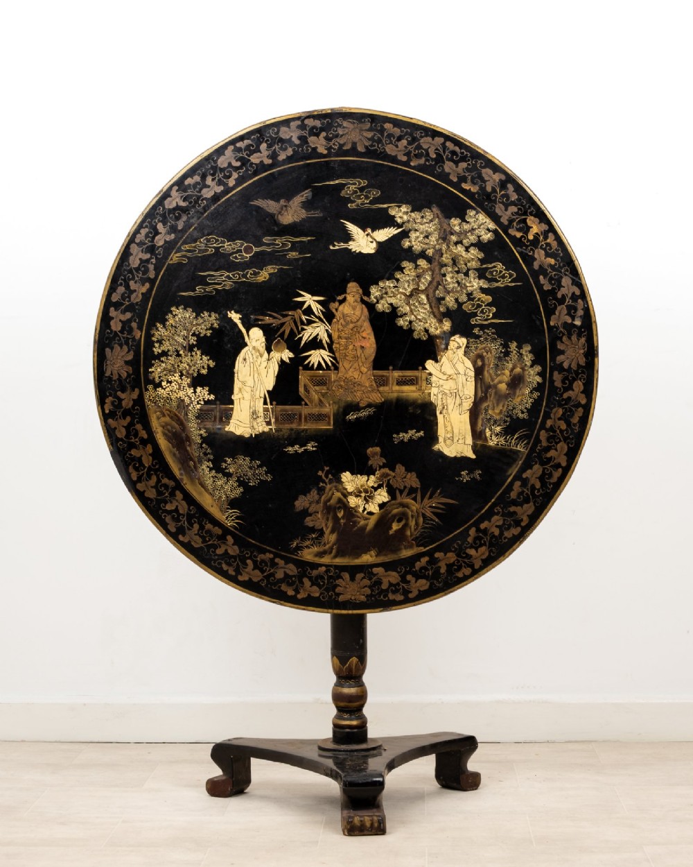 A circular black lacquer tilt-top table decorated with gilded Chinese figures, flora, and fauna on a turned pedestal tripod base.