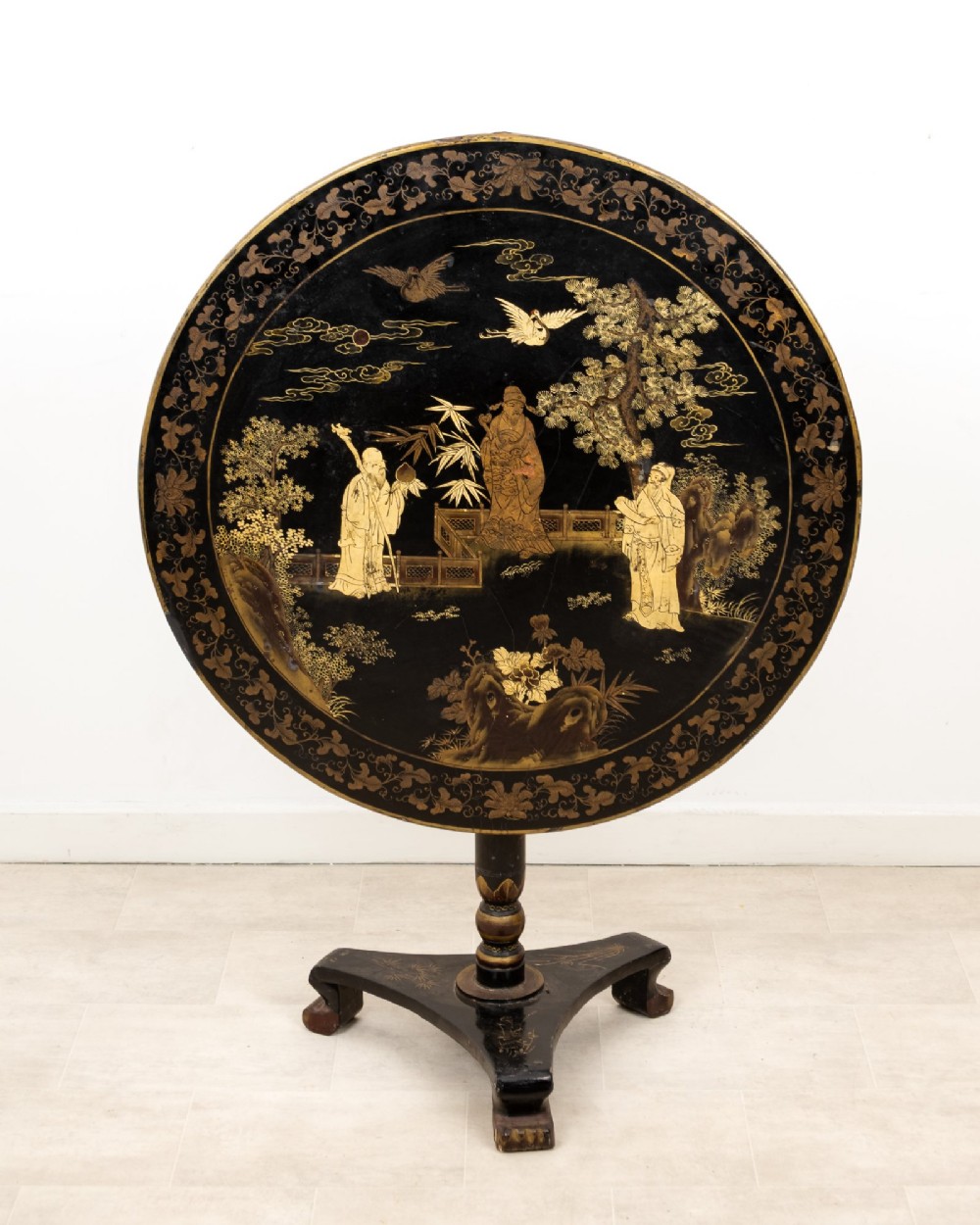 A circular black lacquer tilt-top table decorated with gilded Chinese figures, flora, and fauna on a turned pedestal tripod base. View II