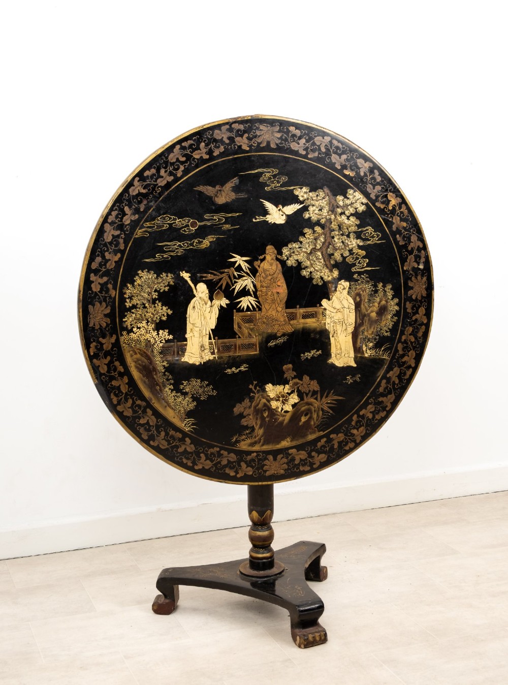 A circular black lacquer tilt-top table decorated with gilded Chinese figures, flora, and fauna on a turned pedestal tripod base. View III