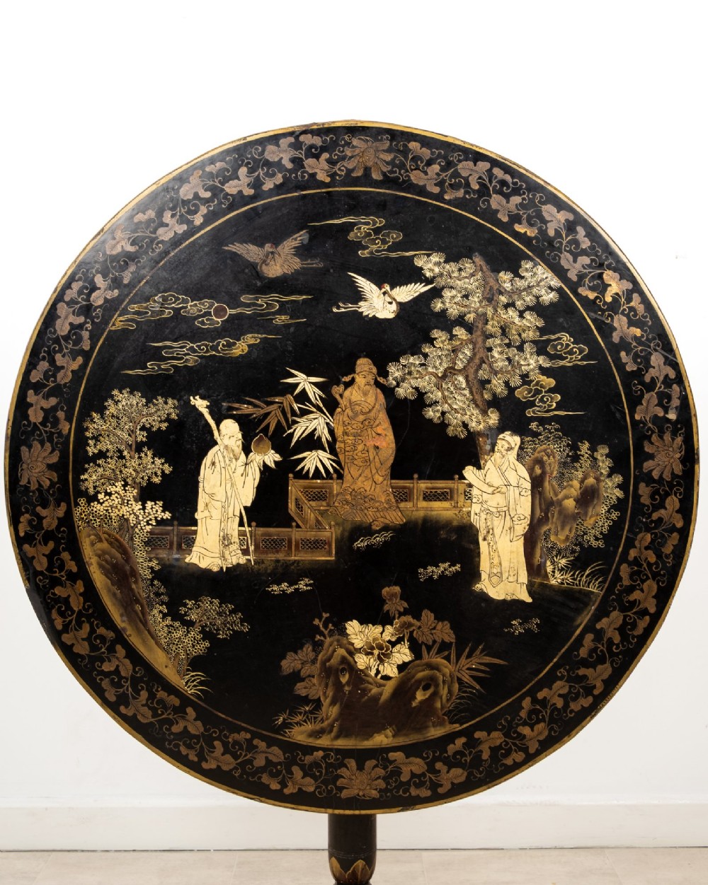 A circular black lacquer tilt-top table decorated with gilded Chinese figures, flora, and fauna on a turned pedestal tripod base. View V