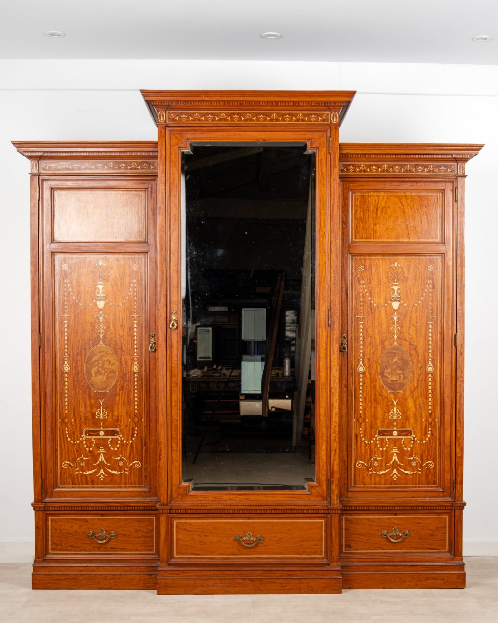 An Edwardian satinwood triple wardrobe with a stepped cornice, central mirror door, side doors decorated with painted classical urns, and three lower drawers.