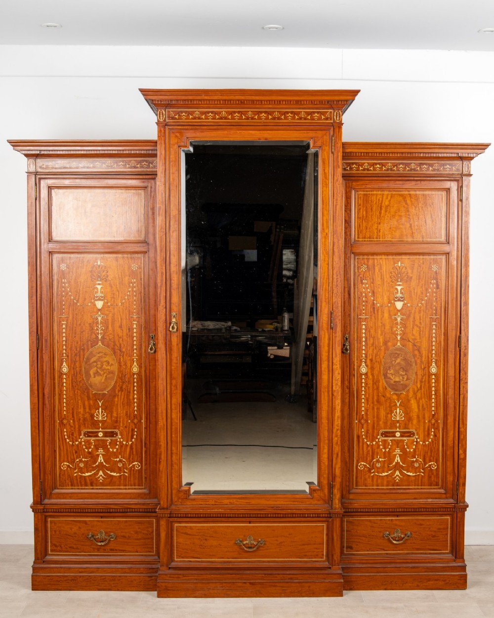 An Edwardian satinwood triple wardrobe with a stepped cornice, central mirror door, side doors decorated with painted classical urns, and three lower drawers. View II