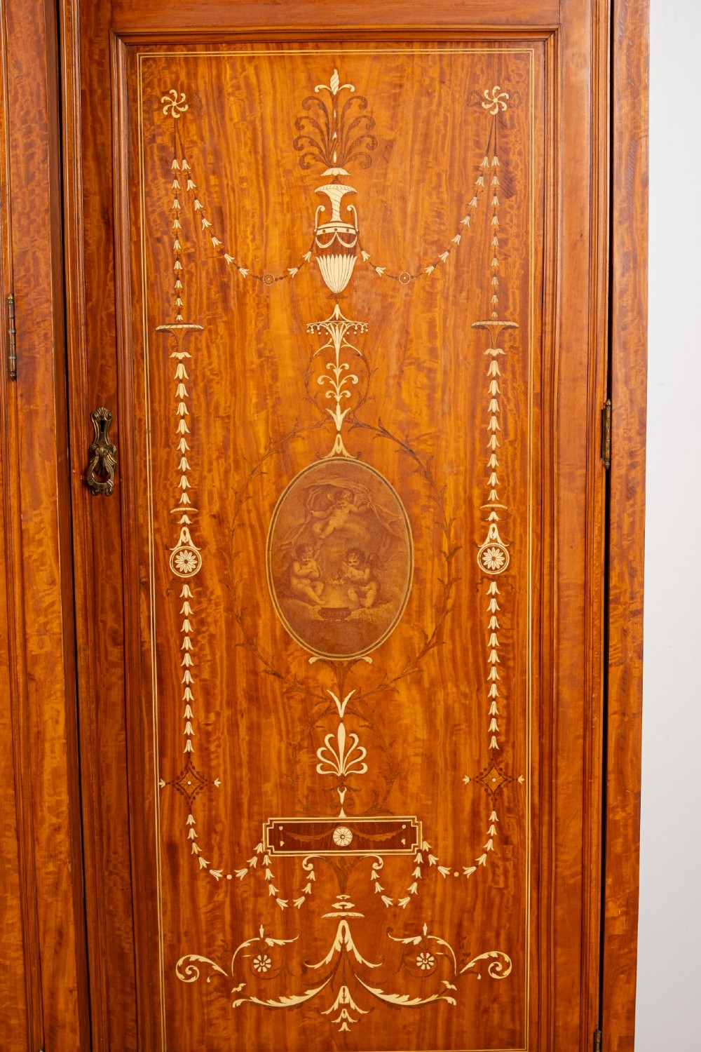 An Edwardian satinwood triple wardrobe with a stepped cornice, central mirror door, side doors decorated with painted classical urns, and three lower drawers. View IV