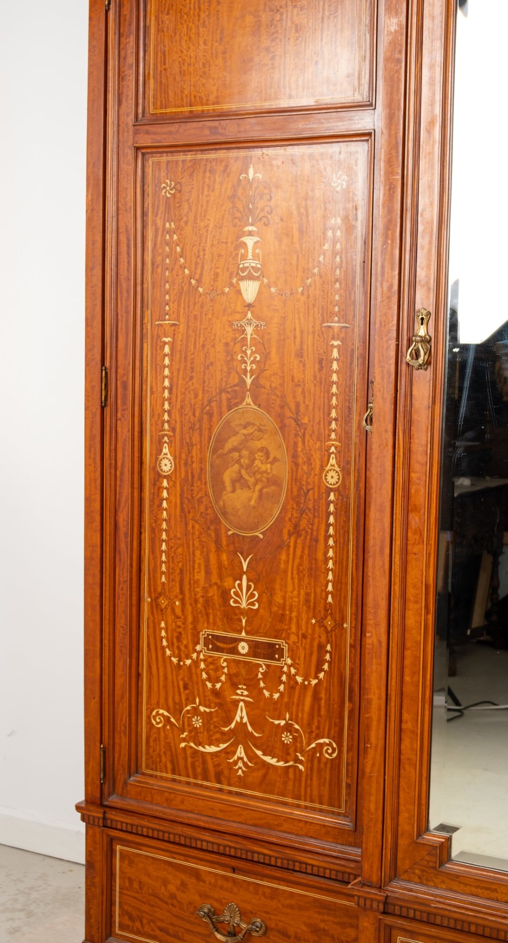 An Edwardian satinwood triple wardrobe with a stepped cornice, central mirror door, side doors decorated with painted classical urns, and three lower drawers. View V