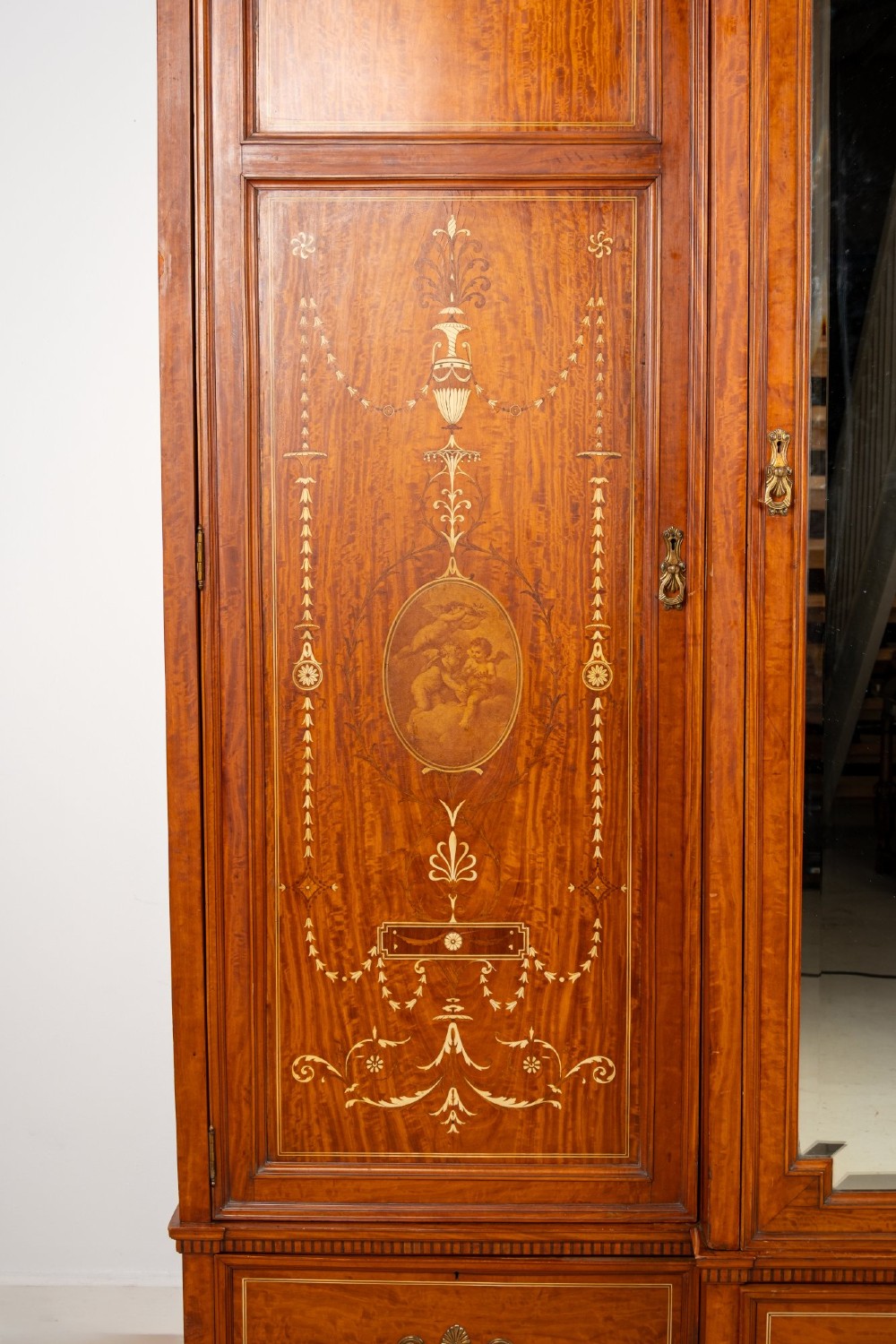 An Edwardian satinwood triple wardrobe with a stepped cornice, central mirror door, side doors decorated with painted classical urns, and three lower drawers. View VI