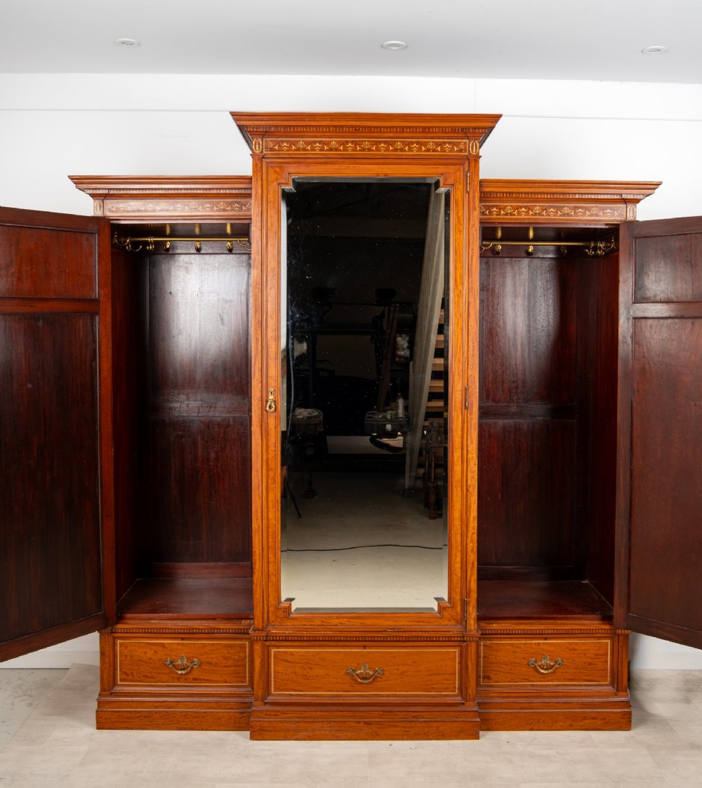 An Edwardian satinwood triple wardrobe with a stepped cornice, central mirror door, side doors decorated with painted classical urns, and three lower drawers. View IX