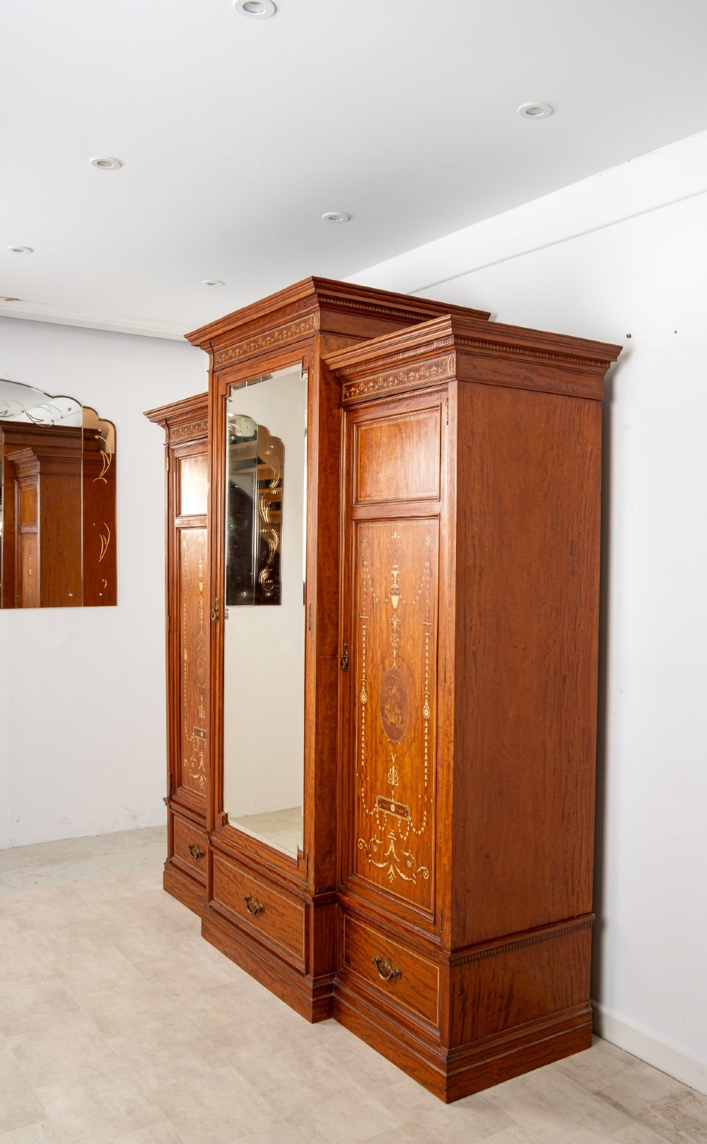 An Edwardian satinwood triple wardrobe with a stepped cornice, central mirror door, side doors decorated with painted classical urns, and three lower drawers. View X
