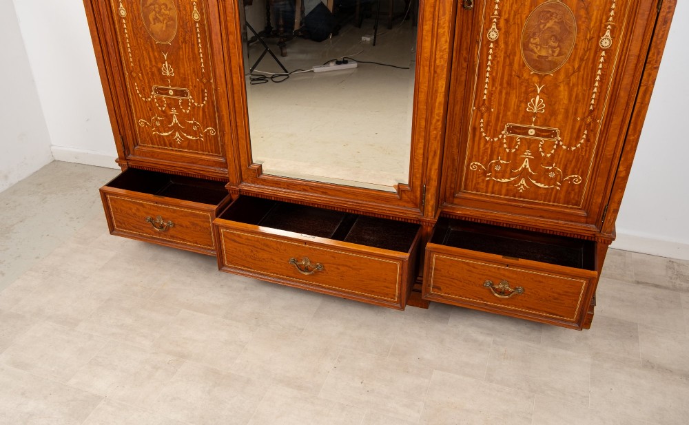 An Edwardian satinwood triple wardrobe with a stepped cornice, central mirror door, side doors decorated with painted classical urns, and three lower drawers. View XI