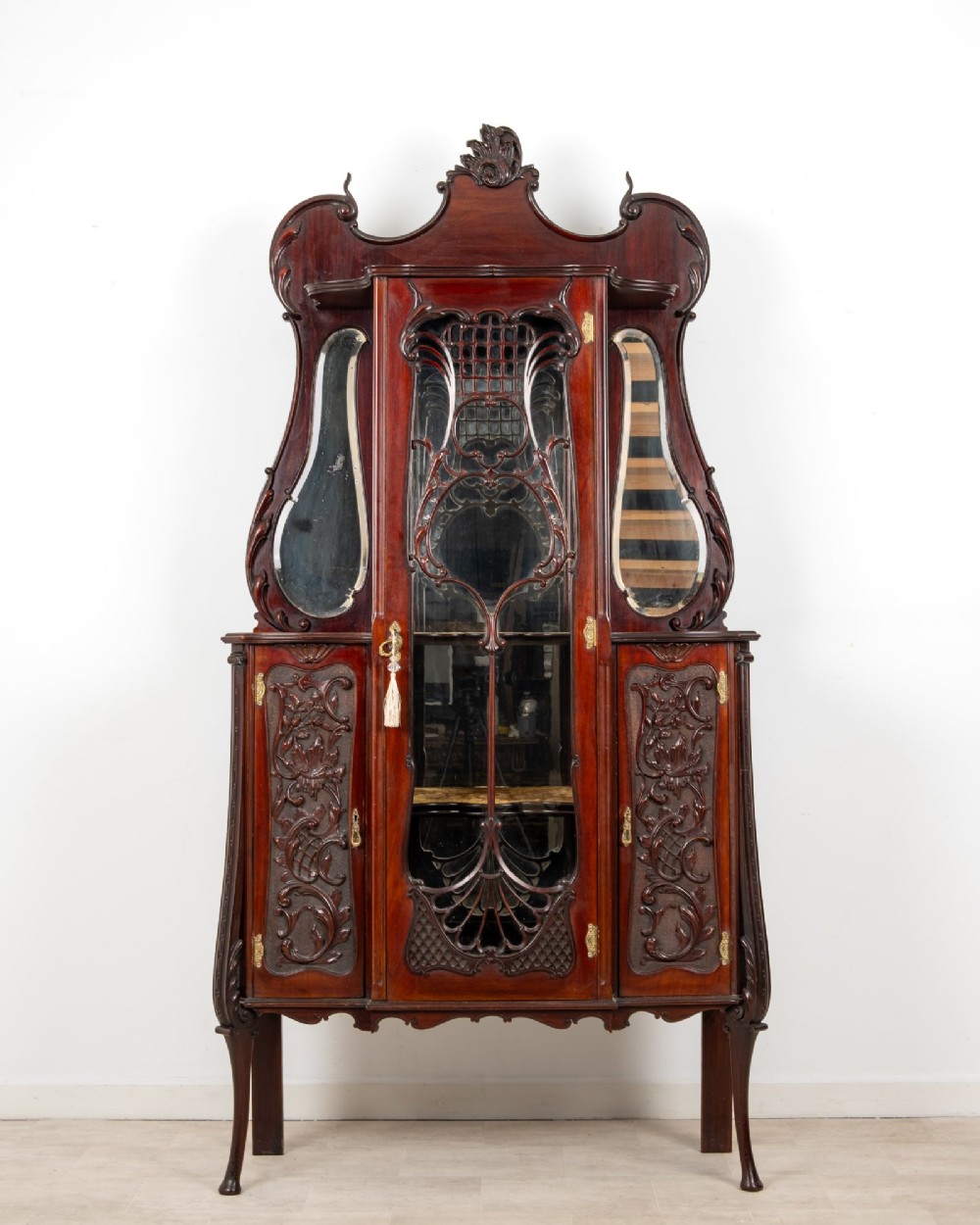 Late Victorian mahogany display cabinet featuring a central glazed door with intricate carved tracery, flanking carved timber doors, side mirror panels, and shaped cresting. View II