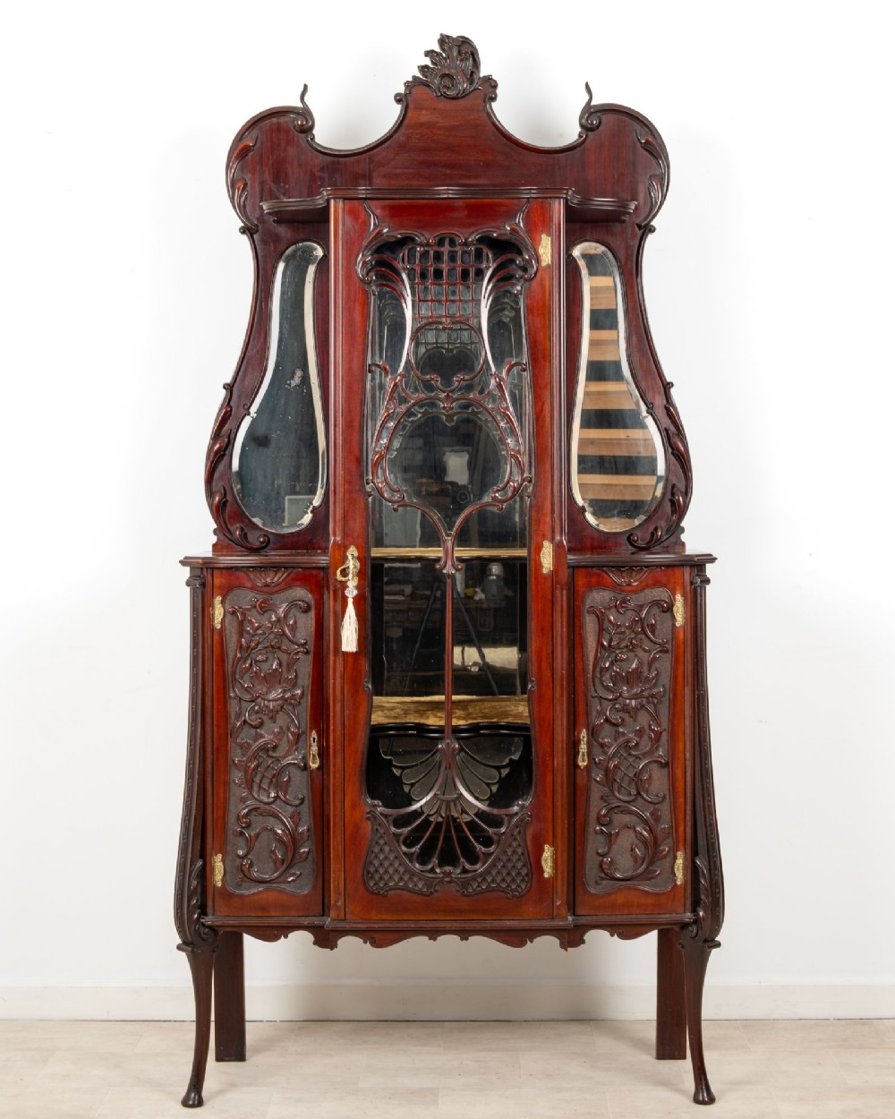 Late Victorian mahogany display cabinet featuring a central glazed door with intricate carved tracery, flanking carved timber doors, side mirror panels, and shaped cresting.