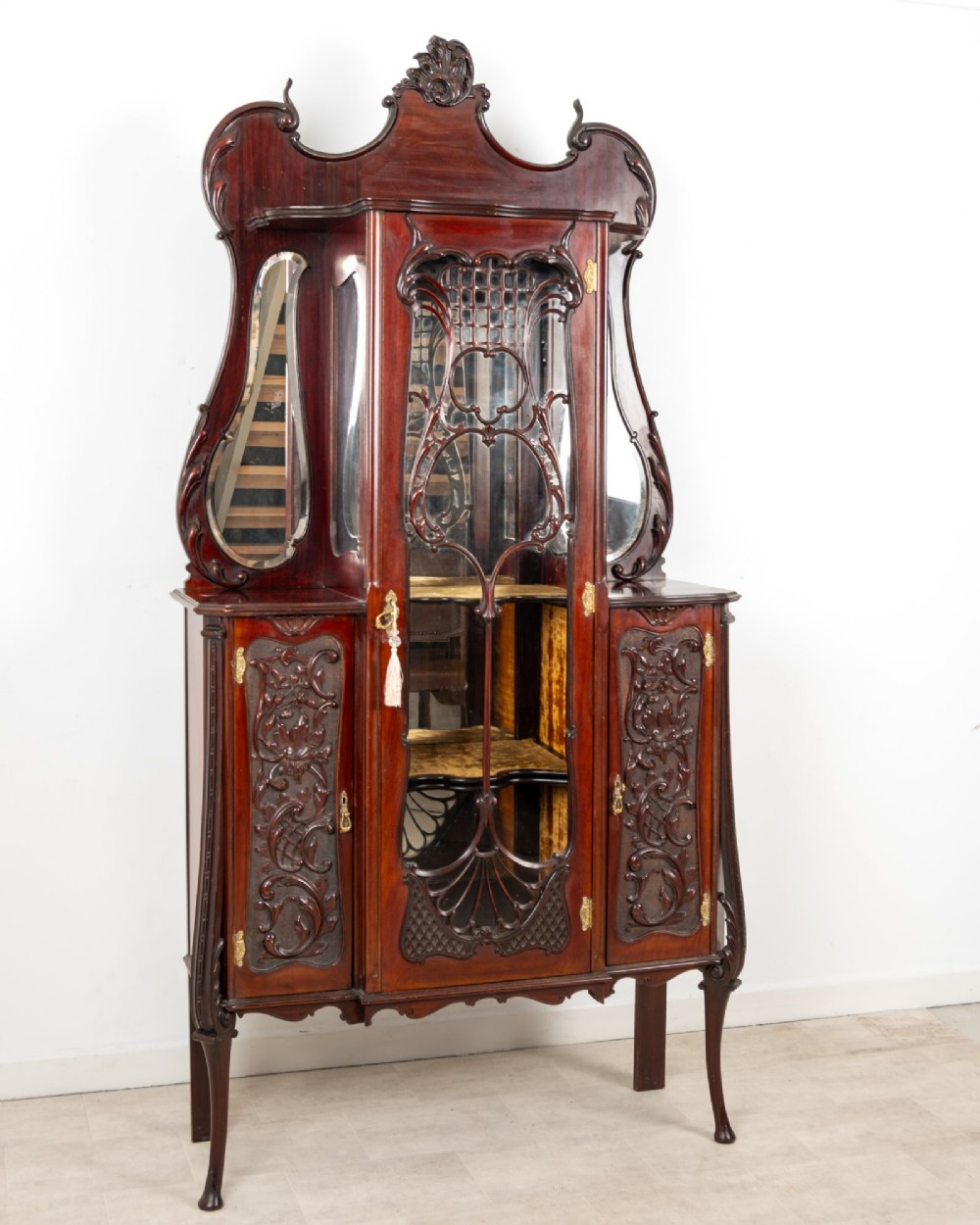 Late Victorian mahogany display cabinet featuring a central glazed door with intricate carved tracery, flanking carved timber doors, side mirror panels, and shaped cresting. View III