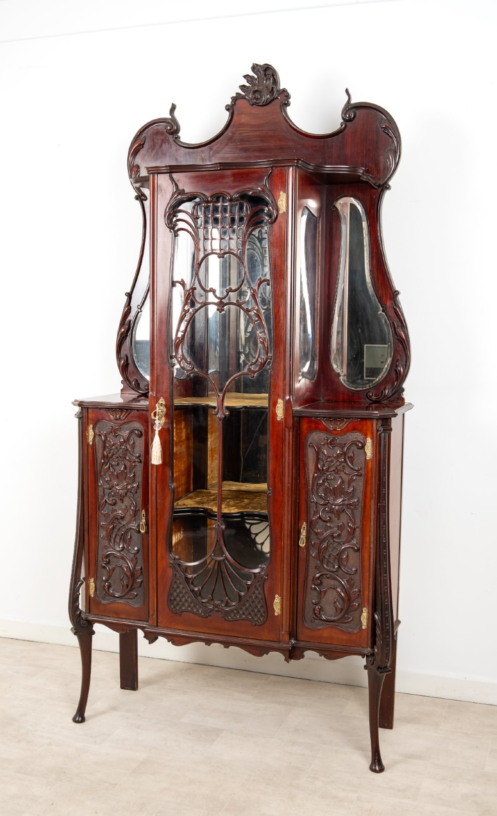 Late Victorian mahogany display cabinet featuring a central glazed door with intricate carved tracery, flanking carved timber doors, side mirror panels, and shaped cresting. View IV