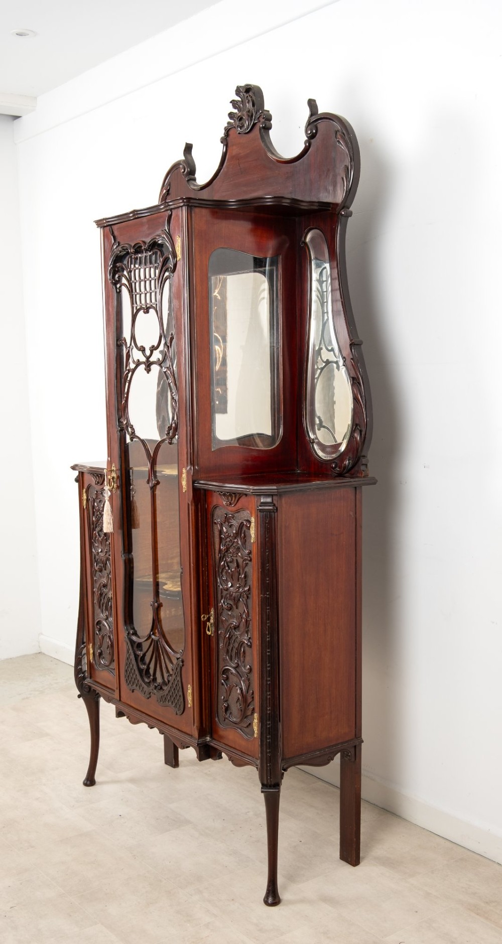 Late Victorian mahogany display cabinet featuring a central glazed door with intricate carved tracery, flanking carved timber doors, side mirror panels, and shaped cresting. View X