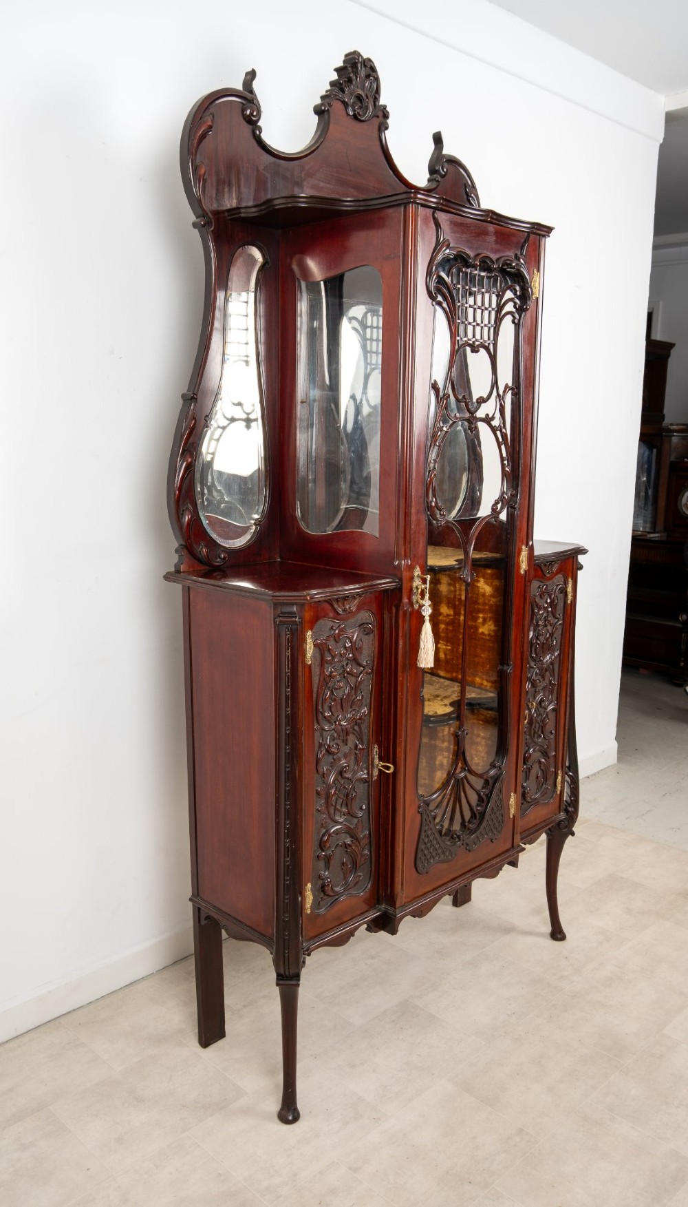 Late Victorian mahogany display cabinet featuring a central glazed door with intricate carved tracery, flanking carved timber doors, side mirror panels, and shaped cresting. View XI