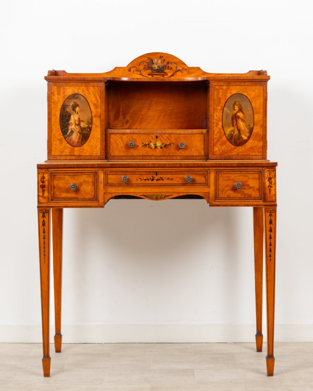 An Edwardian hand-painted satinwood writing desk featuring a raised central cupboard superstructure decorated with painted oval portraits, floral swags, and square tapered legs.