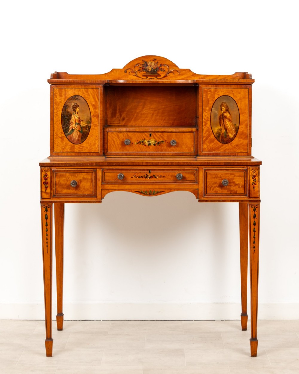 An Edwardian hand-painted satinwood writing desk featuring a raised central cupboard superstructure decorated with painted oval portraits, floral swags, and square tapered legs. View II