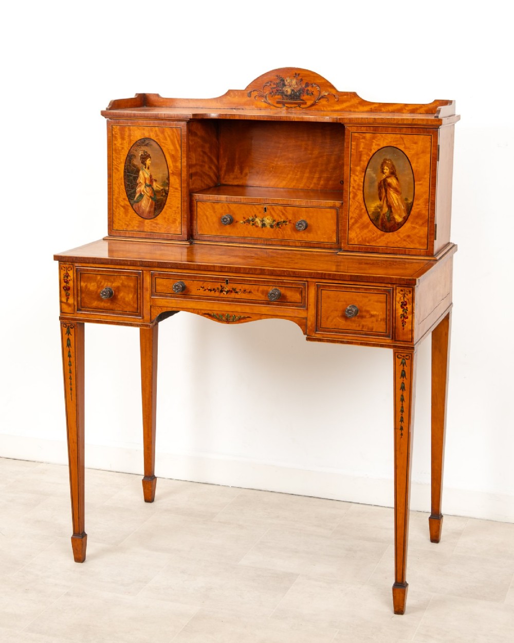 An Edwardian hand-painted satinwood writing desk featuring a raised central cupboard superstructure decorated with painted oval portraits, floral swags, and square tapered legs. View IV