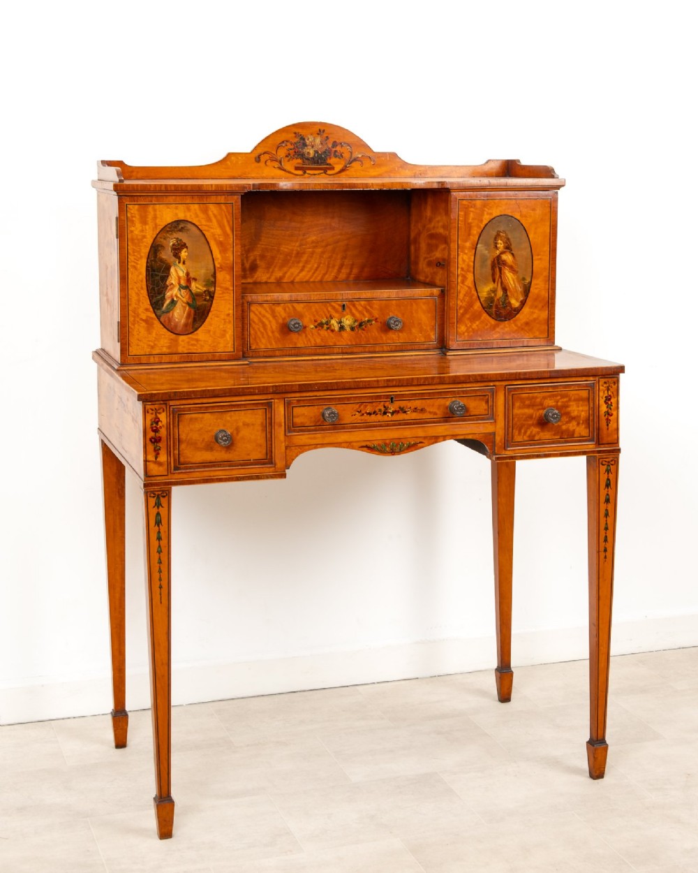 An Edwardian hand-painted satinwood writing desk featuring a raised central cupboard superstructure decorated with painted oval portraits, floral swags, and square tapered legs. View V