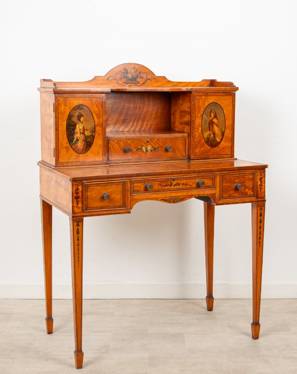 An Edwardian hand-painted satinwood writing desk featuring a raised central cupboard superstructure decorated with painted oval portraits, floral swags, and square tapered legs. View X