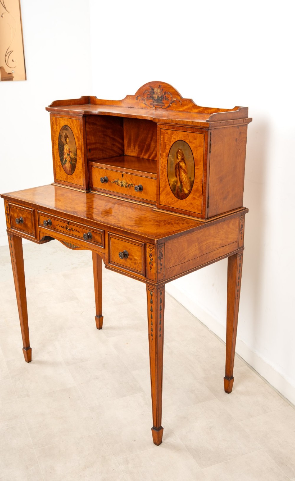 An Edwardian hand-painted satinwood writing desk featuring a raised central cupboard superstructure decorated with painted oval portraits, floral swags, and square tapered legs. View XI