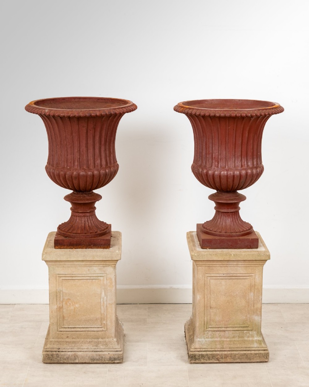 A pair of 19th-century fluted cast iron Campana urns with rust-red finish, elevated on square carved beige stone pedestals. View II