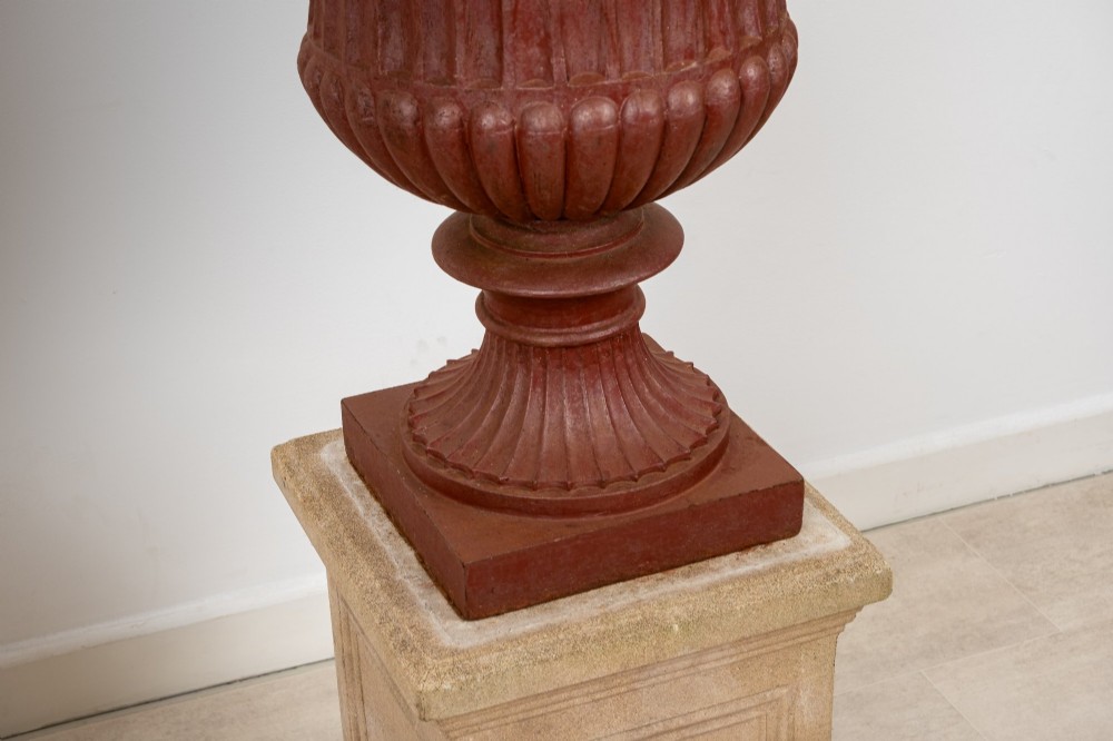 A pair of 19th-century fluted cast iron Campana urns with rust-red finish, elevated on square carved beige stone pedestals. View V