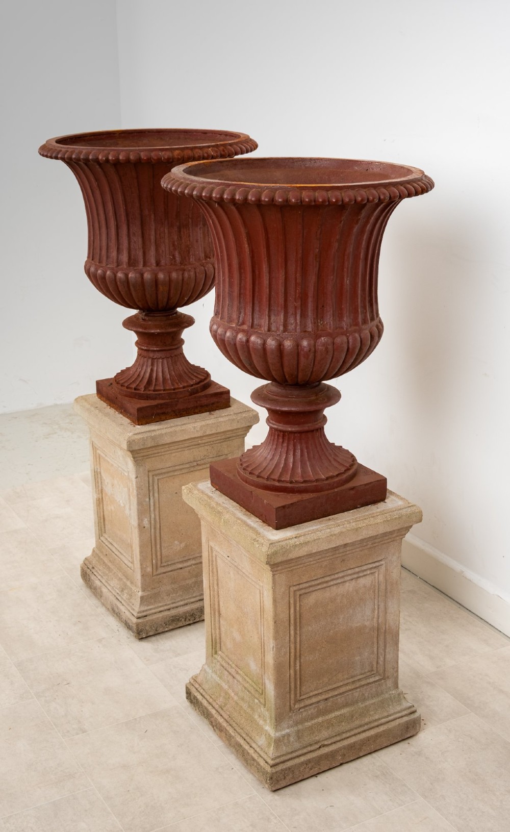 A pair of 19th-century fluted cast iron Campana urns with rust-red finish, elevated on square carved beige stone pedestals. View VI