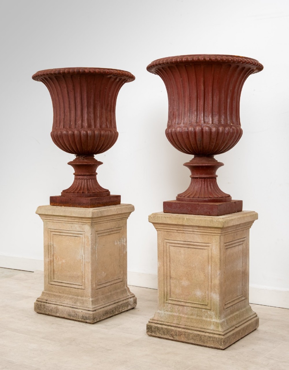 A pair of 19th-century fluted cast iron Campana urns with rust-red finish, elevated on square carved beige stone pedestals. View VII