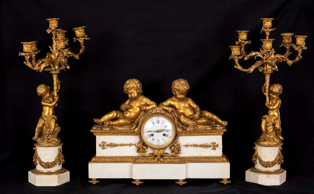 A 19th-century French clock garniture featuring a central white marble and gilt bronze clock topped with two reclining cherub figures, flanked by a pair of matching five-light gilt bronze candelabra supported by cherubs on marble bases.