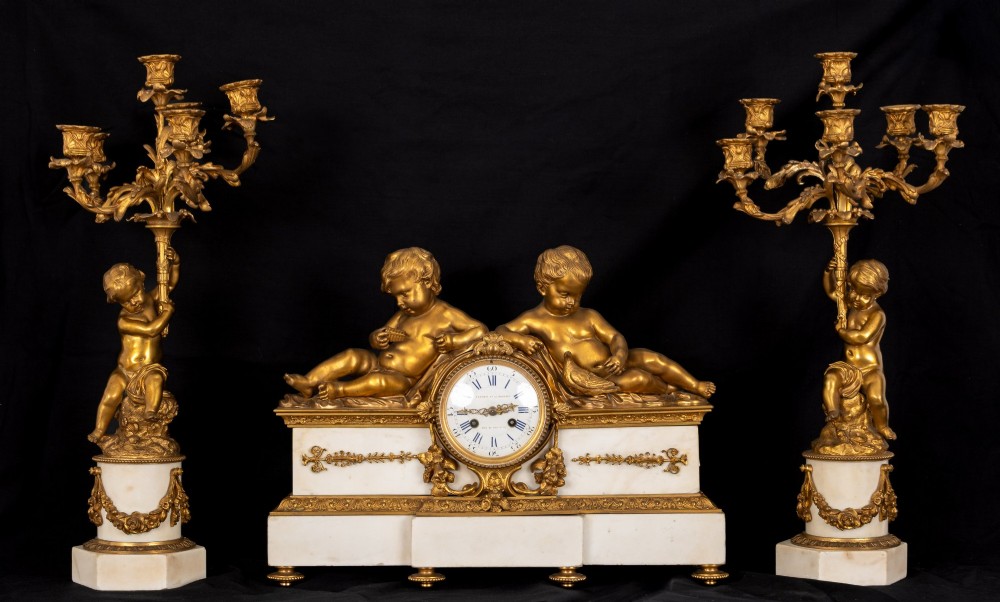 A 19th-century French clock garniture featuring a central white marble and gilt bronze clock topped with two reclining cherub figures, flanked by a pair of matching five-light gilt bronze candelabra supported by cherubs on marble bases. View II