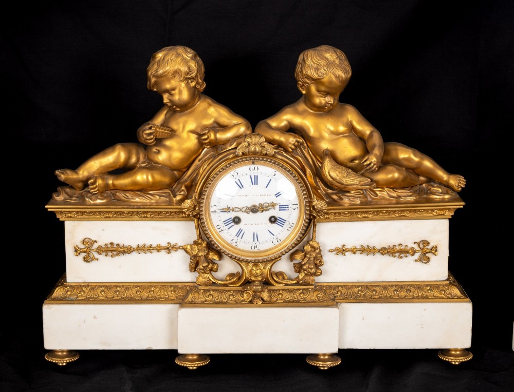 A 19th-century French clock garniture featuring a central white marble and gilt bronze clock topped with two reclining cherub figures, flanked by a pair of matching five-light gilt bronze candelabra supported by cherubs on marble bases. View V