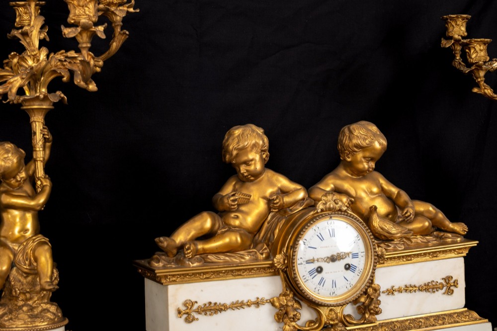 A 19th-century French clock garniture featuring a central white marble and gilt bronze clock topped with two reclining cherub figures, flanked by a pair of matching five-light gilt bronze candelabra supported by cherubs on marble bases. View VI